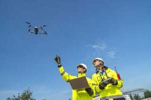Two engineers in high visibility jackets and helmets observing a drone flying over a construction site, with machinery and modular buildings, highlighting technology and site monitoring.