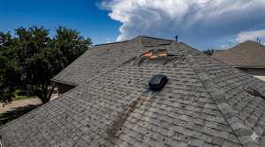 A house roof with missing and damaged shingles, a roof vent, and visible storm damage under a partly cloudy sky.